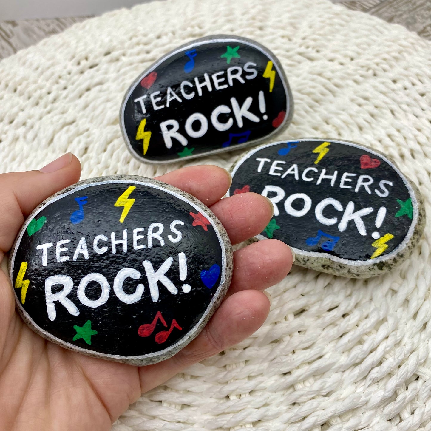 3 stones painted black with rock and roll symbols painted in bright colours and the words "Teachers Rock!" in white