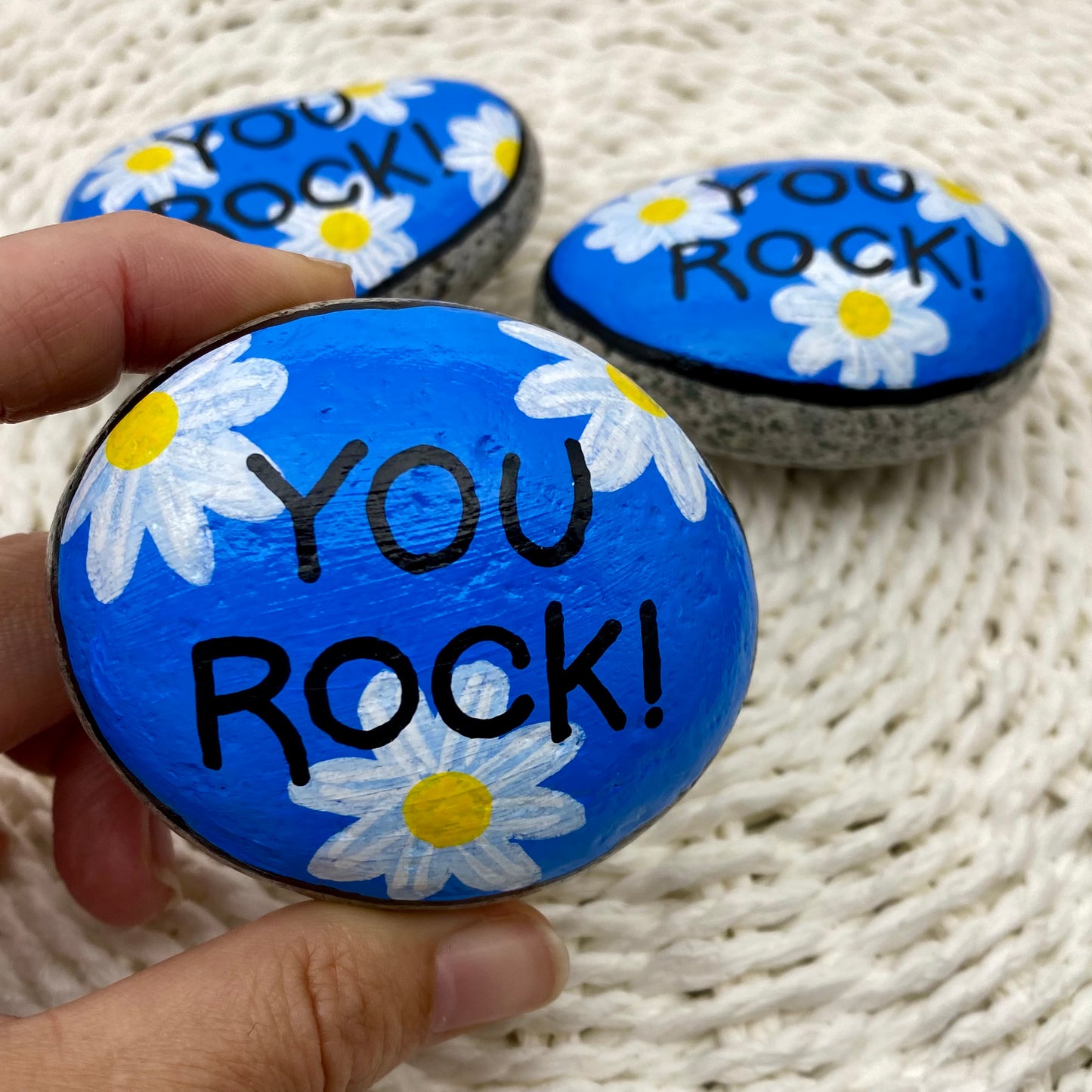 3 rocks painted blue with daisies and the words "You Rock!" painted over the top.