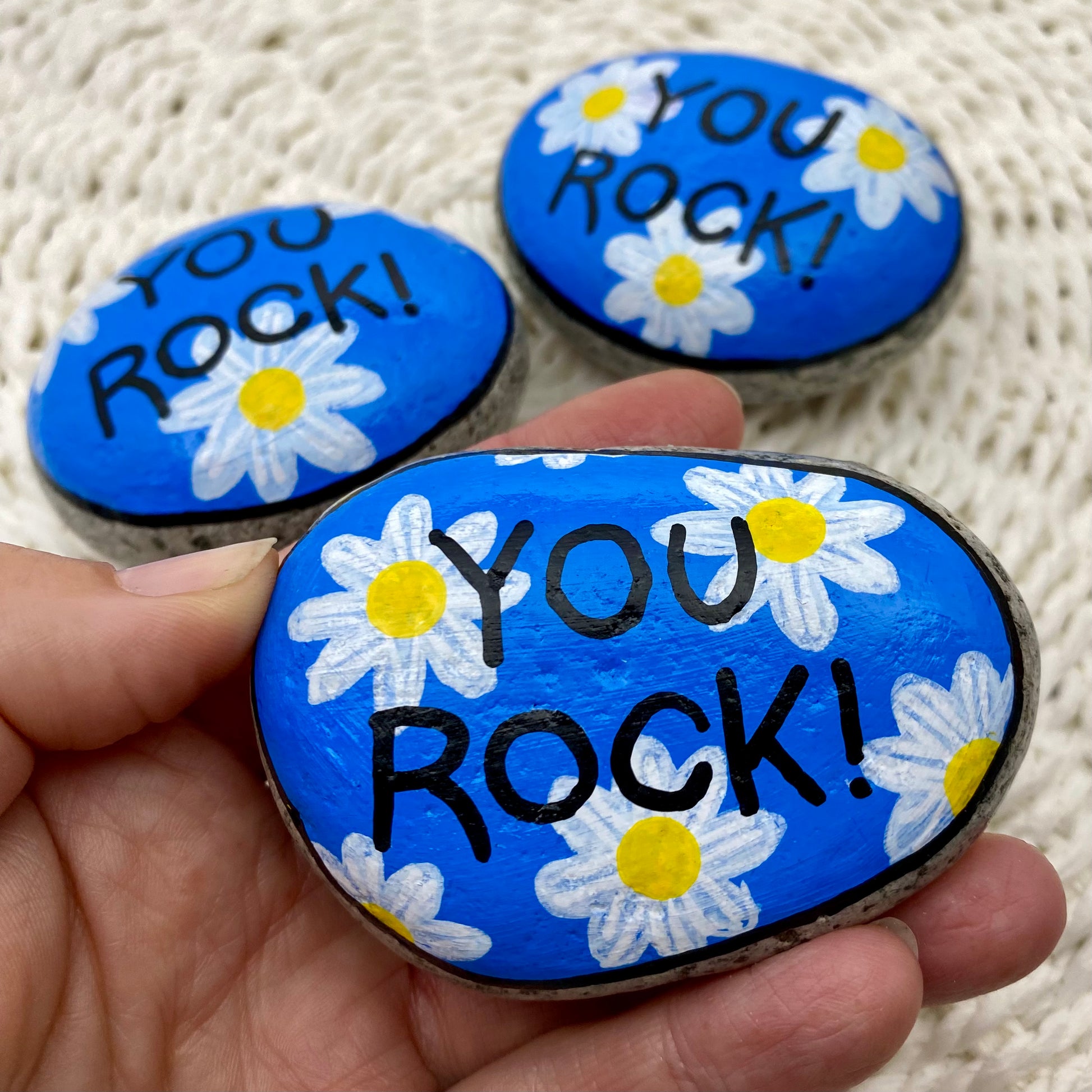 3 rocks painted blue with daisies and the words "You Rock!" painted over the top.