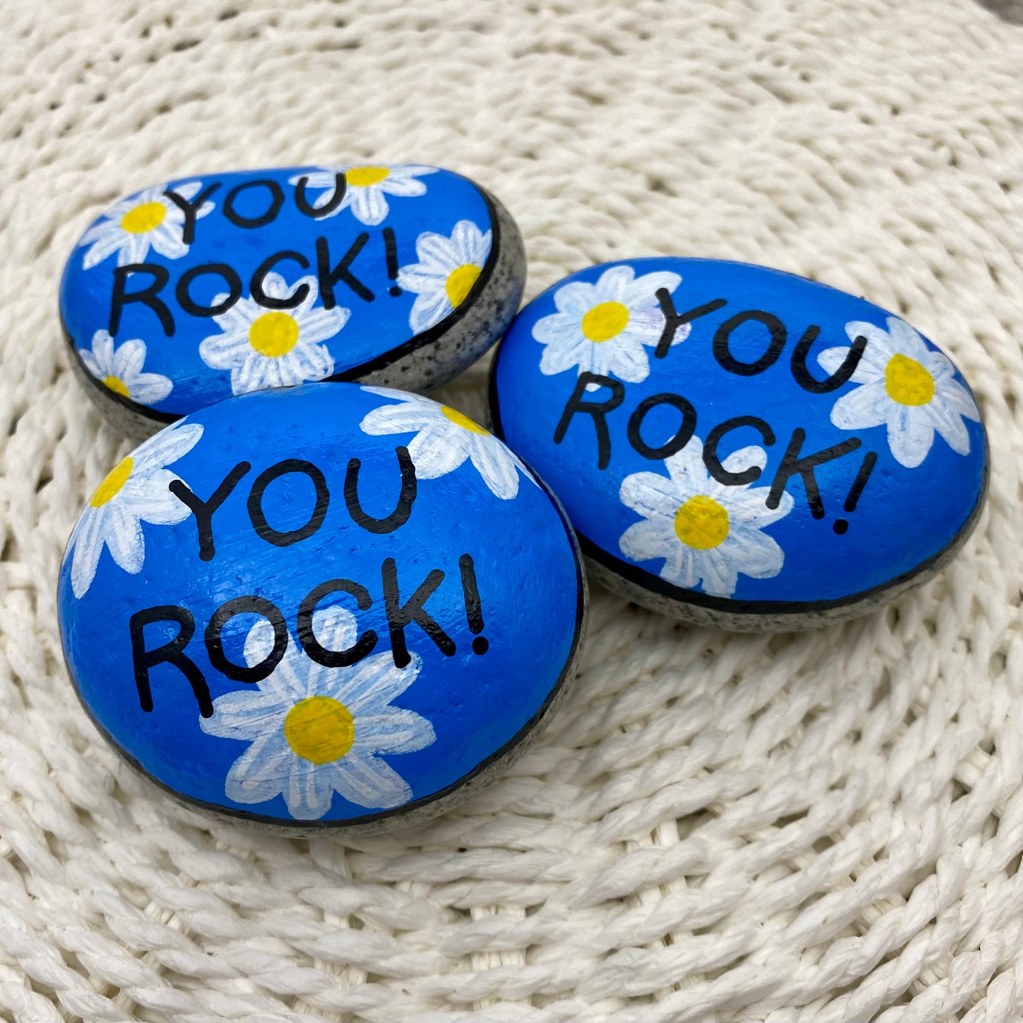 3 rocks painted blue with daisies and the words "You Rock!" painted over the top.