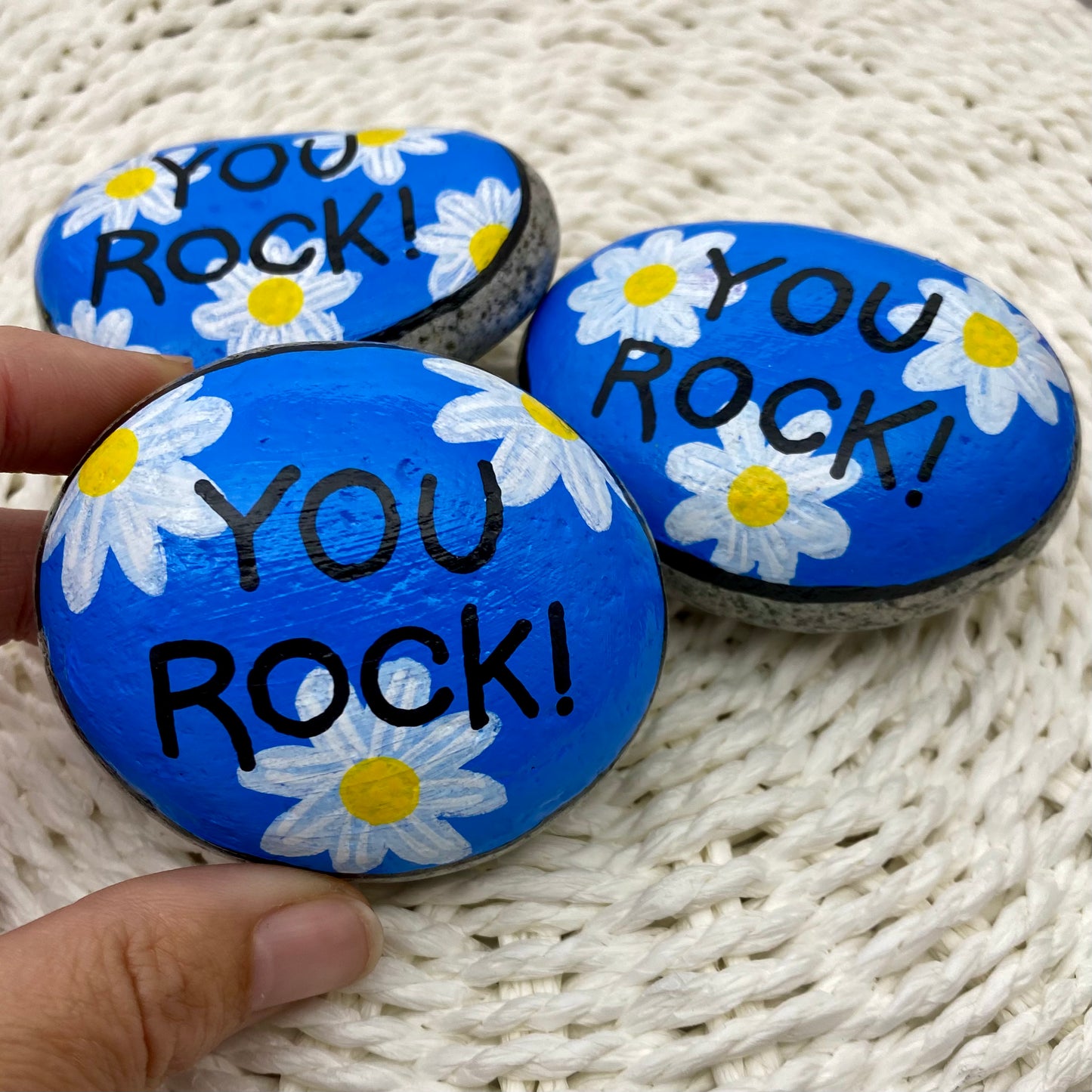 3 rocks painted blue with daisies and the words "You Rock!" painted over the top.