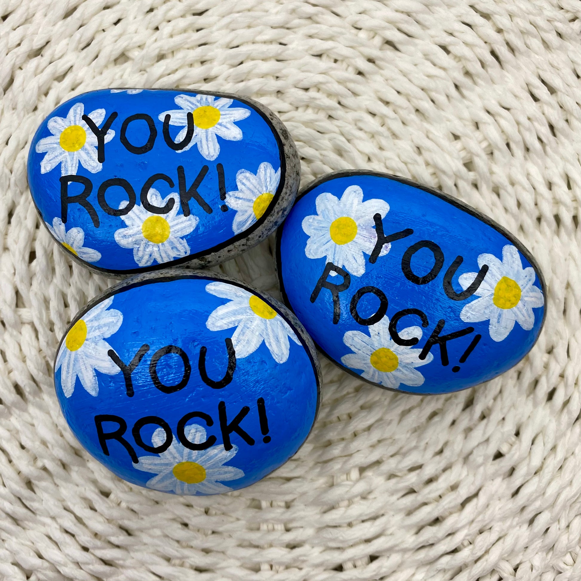 3 rocks painted blue with daisies and the words "You Rock!" painted over the top.