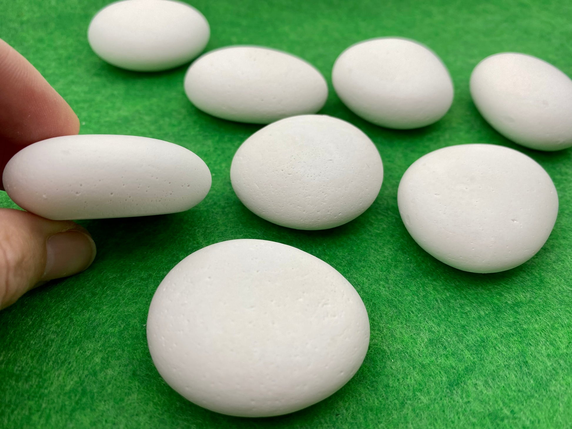 8 small white plaster pebbles of different shapes and sizes, one being held by a hand showing the side of it