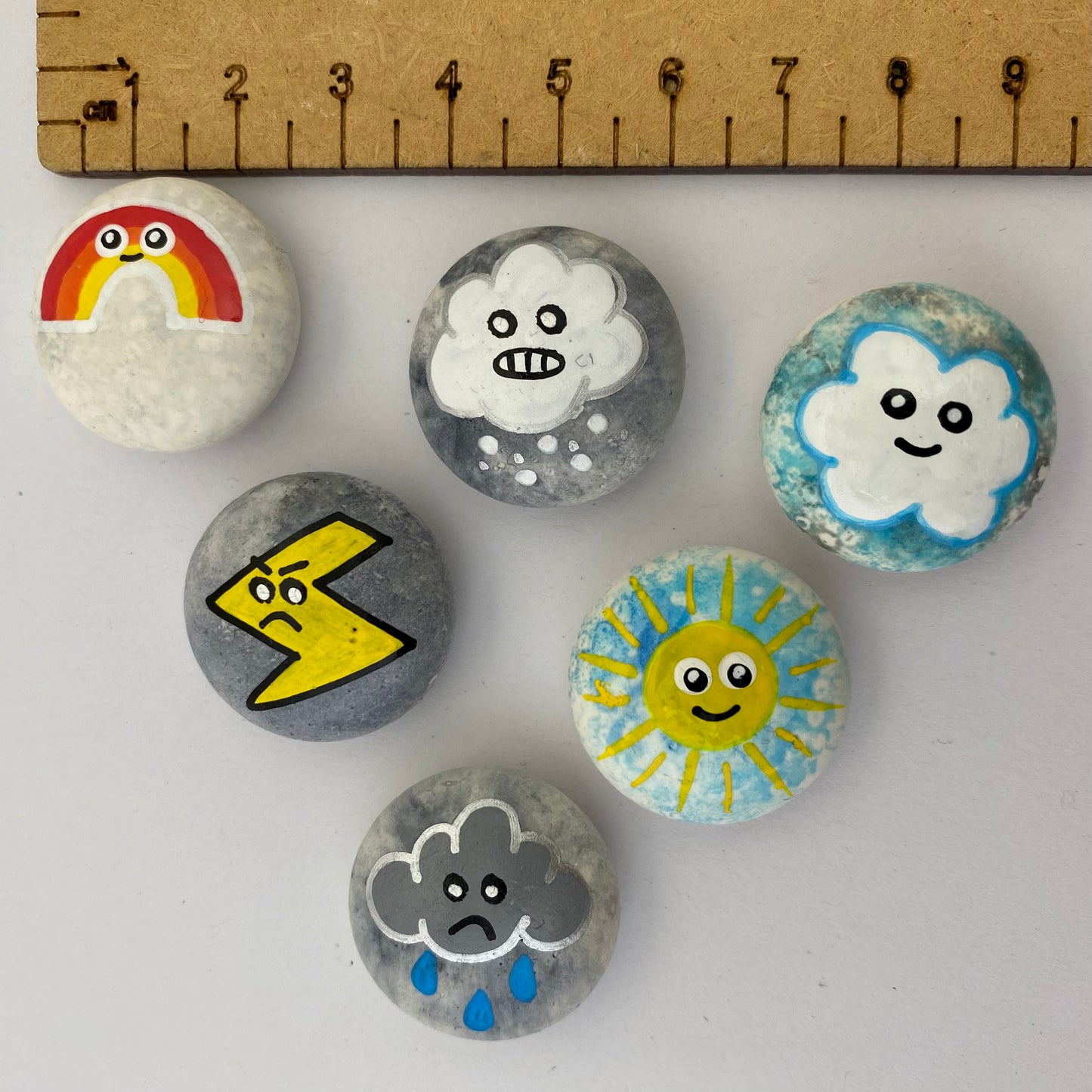 6 small hand painted weather themed pebbles next to a ruler