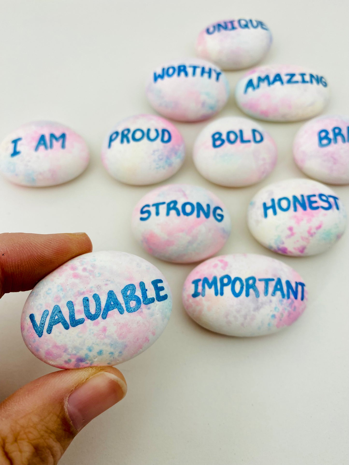 Hand painted affirmation pebbles, one being held closer to the camera with the word Valuable