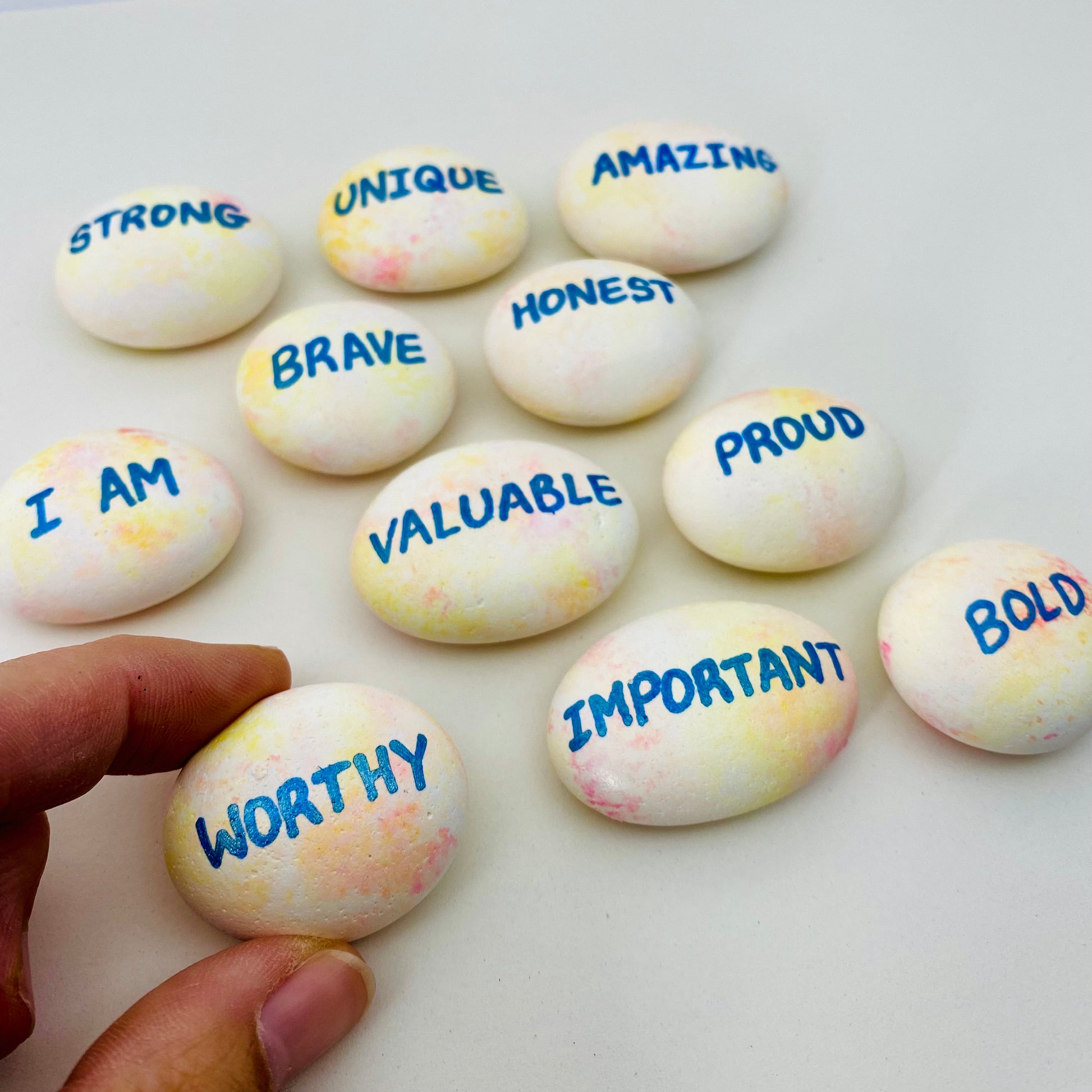 Hand painted affirmation pebbles, one being held closer to the camera with the word Worthy
