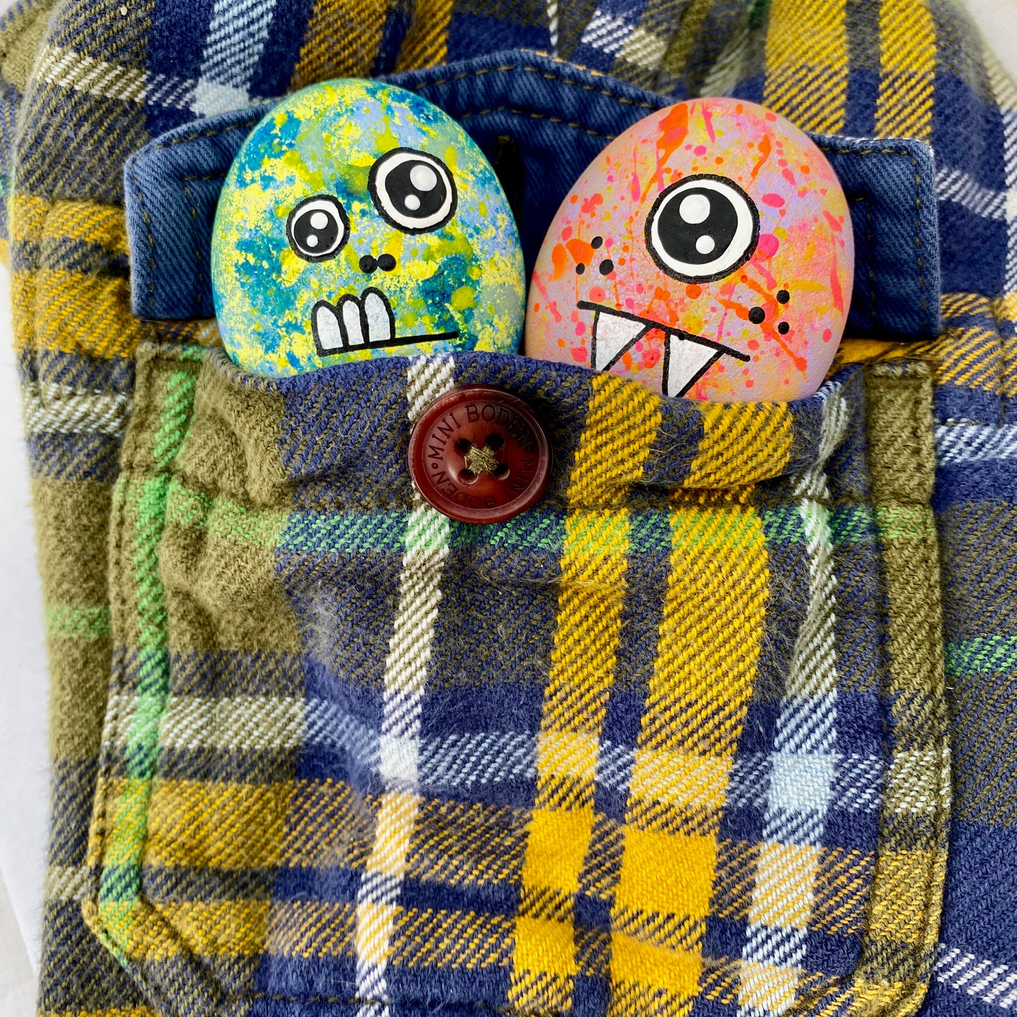 2 hand painted monster pebbles, one with 2 eyes called Flomp, and one with 1 eye called Blerp poking out of a pocket