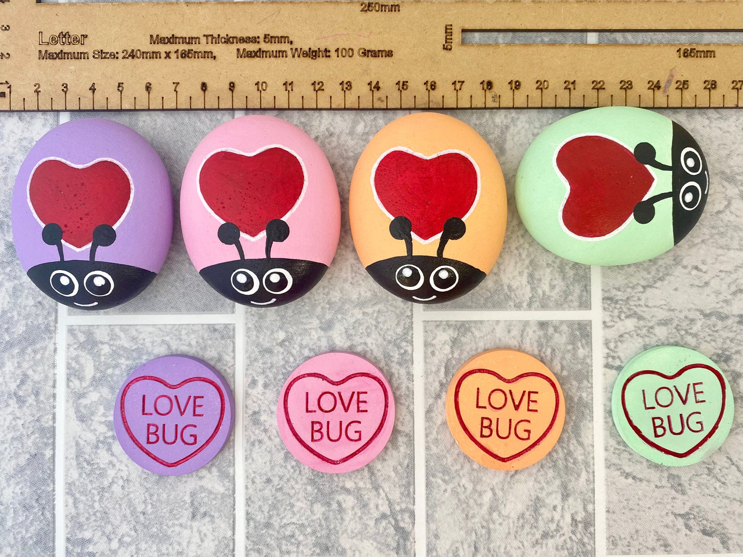 4 Hand painted Pastel Heart Bug Pebbles and Tokens with the words Love Bug inscribed on next to a ruler