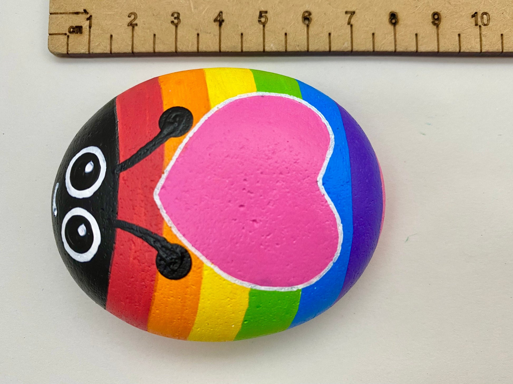Hand painted Rainbow Bug with Heart Pebble next to a ruler